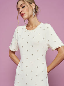 A woman models the Social Queen Beaded Knit Pleated Dress, standing against a pink background and looking to the side with her hands behind her back.