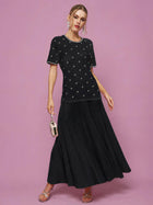 A woman poses against a pink background wearing the Beaded Knit Pleated Dress by Social Queen, styled with silver heels, statement earrings, and a small metallic bag.