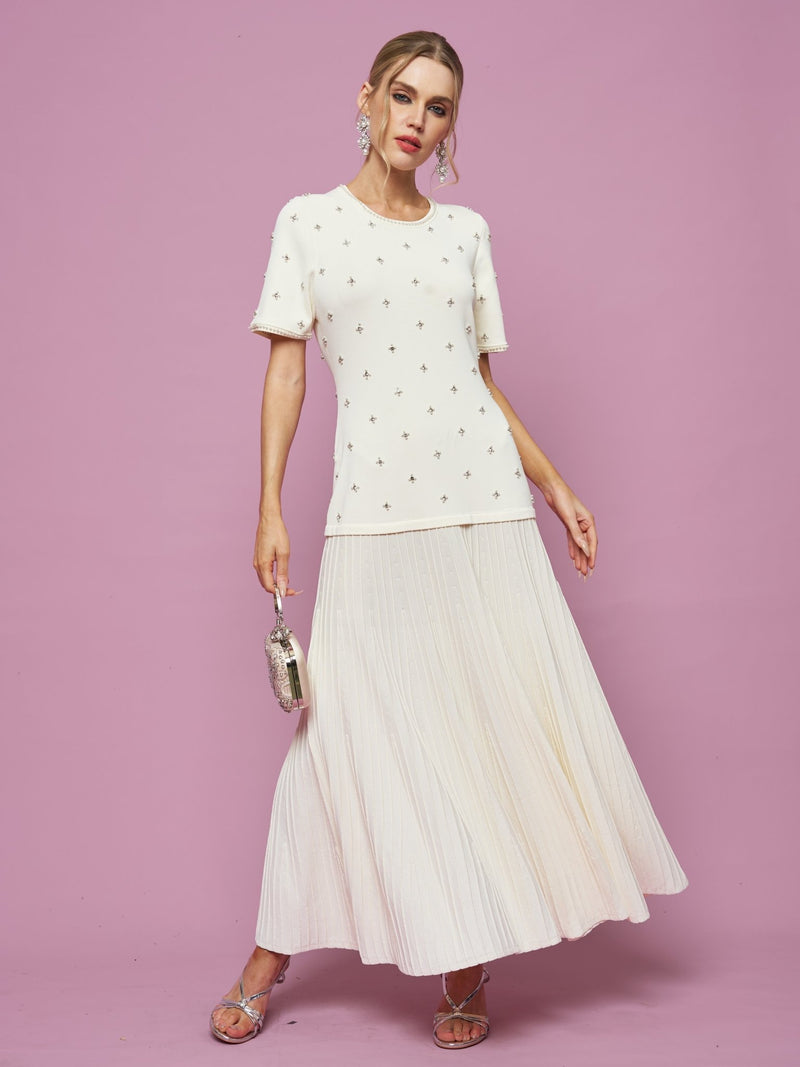 A woman stands against a pink background wearing the Social Queen Beaded Knit Pleated Dress, paired with silver sandals and a small silver clutch.