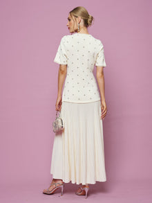 A woman faces away against a pink backdrop, wearing Social Queen’s Beaded Knit Pleated Dress that highlights beaded details and pleats. She pairs it with strappy heels, a small purse, and her hair in a bun.