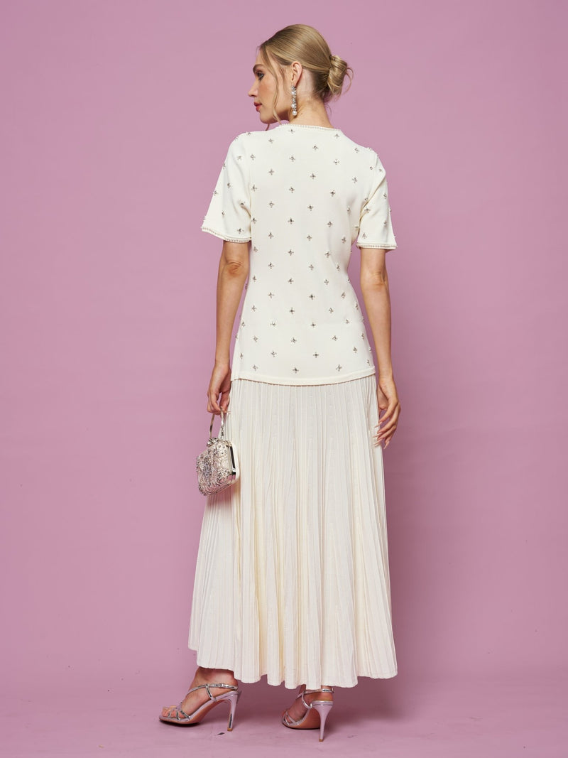 A woman faces away against a pink backdrop, wearing Social Queen’s Beaded Knit Pleated Dress that highlights beaded details and pleats. She pairs it with strappy heels, a small purse, and her hair in a bun.
