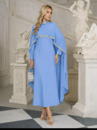 A woman stands indoors near classical statues and a staircase, wearing the Social Queen Beaded Mock-Neck Cape Dress in light blue and holding a small silver handbag.
