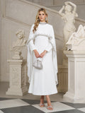 A woman stands indoors near classical statues wearing the Social Queen Beaded Mock-Neck Cape Dress, paired with a silver handbag and heeled sandals.
