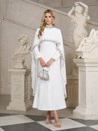 A woman stands indoors near classical statues wearing the Social Queen Beaded Mock-Neck Cape Dress, paired with a silver handbag and heeled sandals.