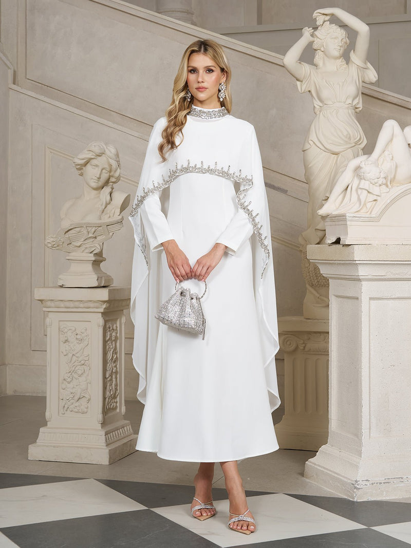 A woman stands indoors near classical statues wearing the Social Queen Beaded Mock-Neck Cape Dress, paired with a silver handbag and heeled sandals.