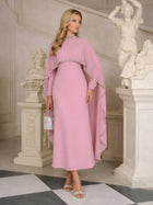 A woman wearing the Social Queen Beaded Mock-Neck Cape Dress in light pink stands indoors by white statues and a staircase, holding a small handbag.