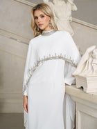 A woman wears the Social Queen Beaded Mock-Neck Cape Dress, featuring silver embellishments, as she stands indoors beside a staircase and classical sculptures.