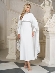 A woman in Social Queen's Beaded Mock-Neck Cape Dress stands indoors by classical statues and a staircase, holding a small silver handbag.