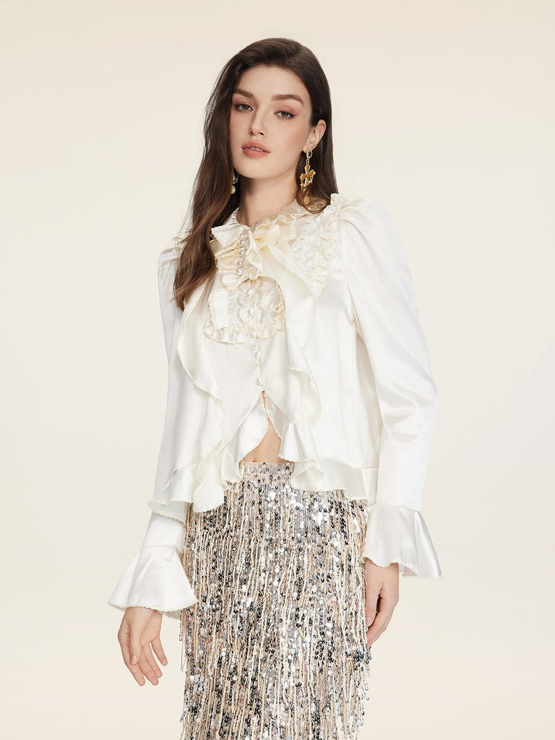 A woman wears the Social Queen Beaded Ruffle Satin Blouse, featuring hand-stitched ruffles and puff sleeves, paired with a silver sequin fringed skirt, posing against a plain light background.