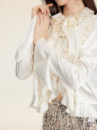 A woman wears the Social Queen Beaded Ruffle Satin Blouse in cream, featuring hand-stitched ruffles and lace, paired with a sequined skirt against a neutral background.