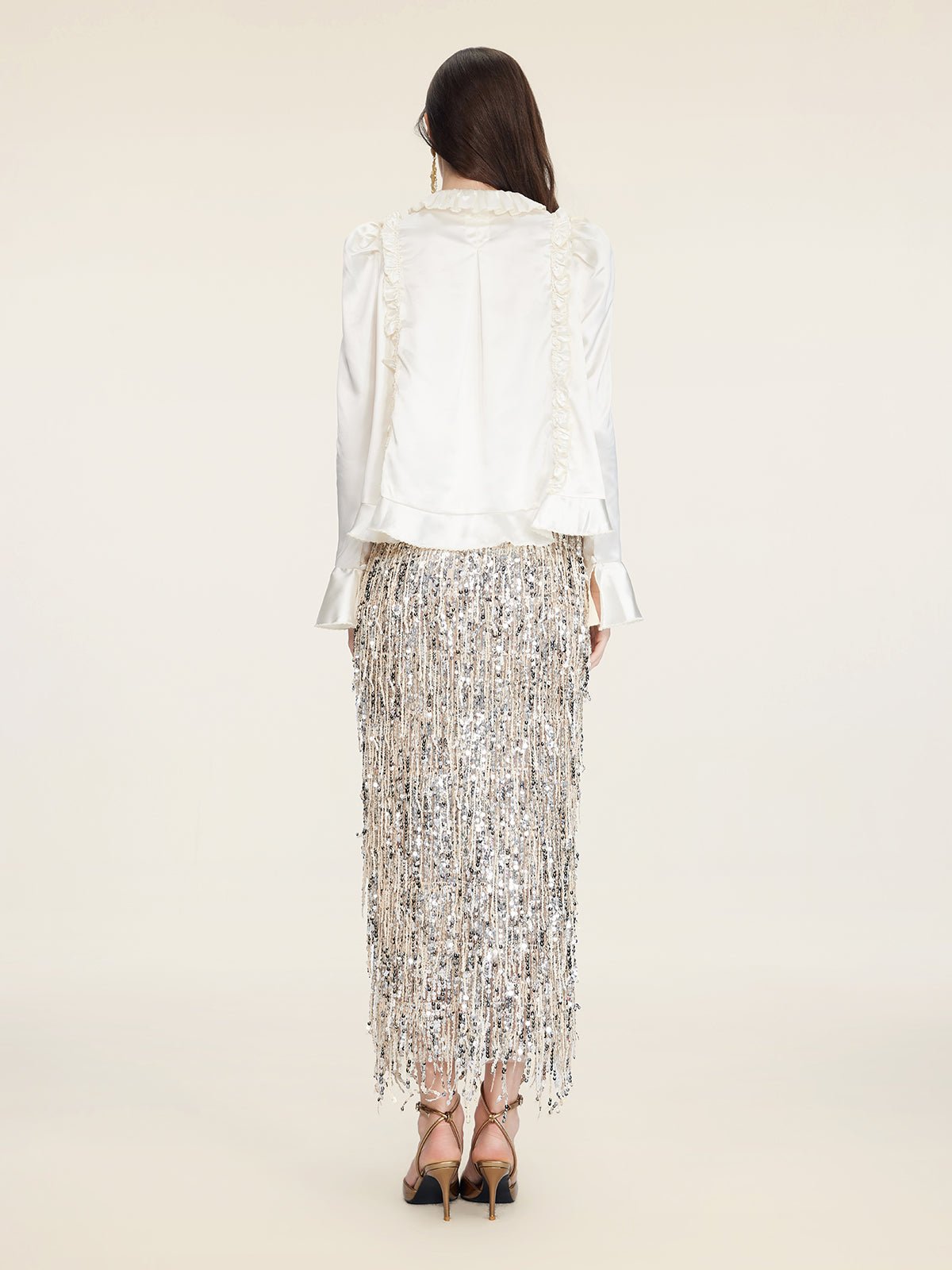 A woman faces away, wearing the Social Queen Beaded Ruffle Satin Blouse with puff sleeves and a silver fringe skirt, paired with high-heeled sandals against a plain light background.