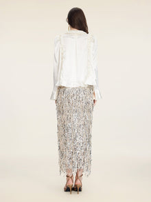 A woman faces away, wearing the Social Queen Beaded Ruffle Satin Blouse with puff sleeves and a silver fringe skirt, paired with high-heeled sandals against a plain light background.