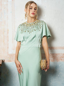 A woman wears the Social Queen Beaded Satin Evening Gown in light green with gold bead accents, paired with a gold clutch and earrings, standing against a textured wall.