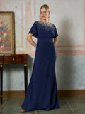 A woman in the Social Queen Beaded Satin Evening Gown—navy blue with short sleeves and beaded neckline—stands indoors by a wooden table and a framed floral painting.