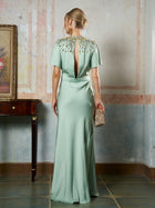 A woman models the Social Queen Beaded Satin Evening Gown in light green, featuring bead embellishment at the neckline and a keyhole back, while holding an embellished clutch.