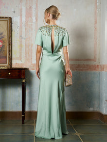 A woman models the Social Queen Beaded Satin Evening Gown in light green, featuring bead embellishment at the neckline and a keyhole back, while holding an embellished clutch.