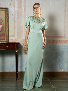 A woman stands indoors wearing the Social Queen Beaded Satin Evening Gown in light green, featuring beaded embellishments on the neckline and holding a small clutch bag.