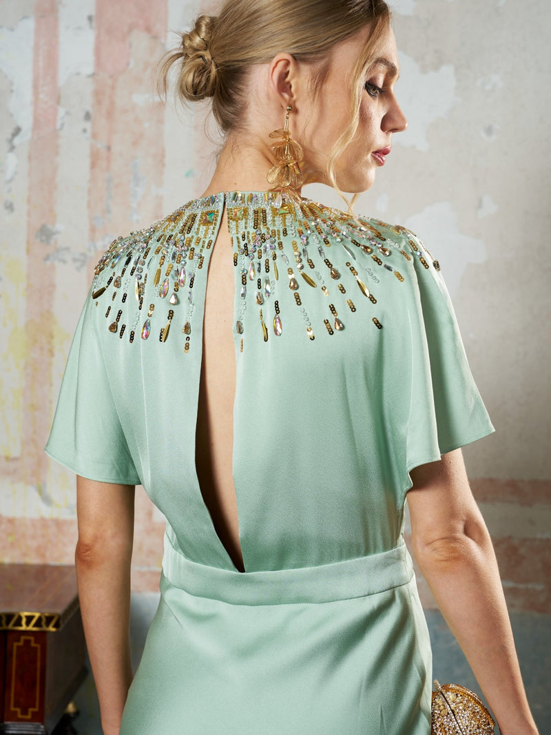 A woman with blonde hair in a low bun wears gold statement earrings and the Social Queen Beaded Satin Evening Gown in light green, featuring bead embellishments, a jeweled neckline, and back keyhole detail as she stands facing away.
