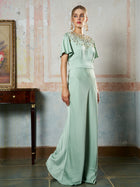 A woman poses indoors by a wooden table and framed painting, wearing the Social Queen Beaded Satin Evening Gown—a floor-length, light green satin dress featuring short sleeves and an ornate beaded neckline.