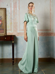 A woman poses indoors by a wooden table and framed painting, wearing the Social Queen Beaded Satin Evening Gown—a floor-length, light green satin dress featuring short sleeves and an ornate beaded neckline.