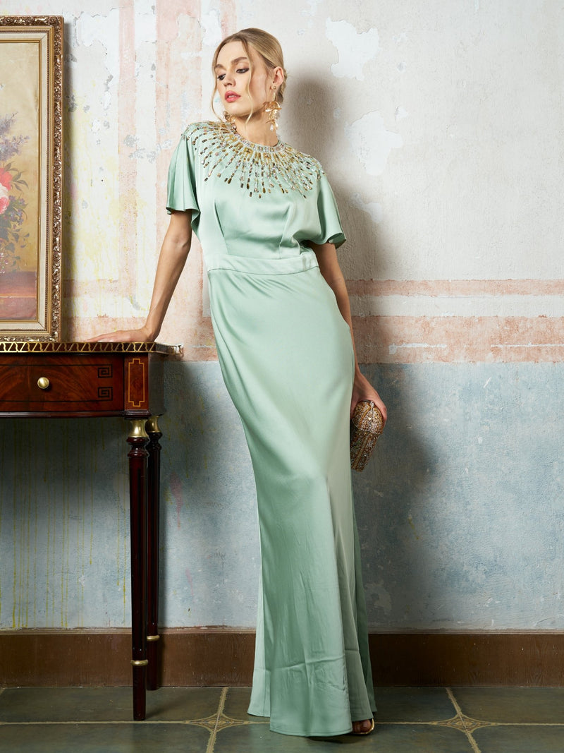 A woman in the Social Queen Beaded Satin Evening Gown, mint green with beaded neckline, holds a clutch while leaning against a wooden table near a painted wall.