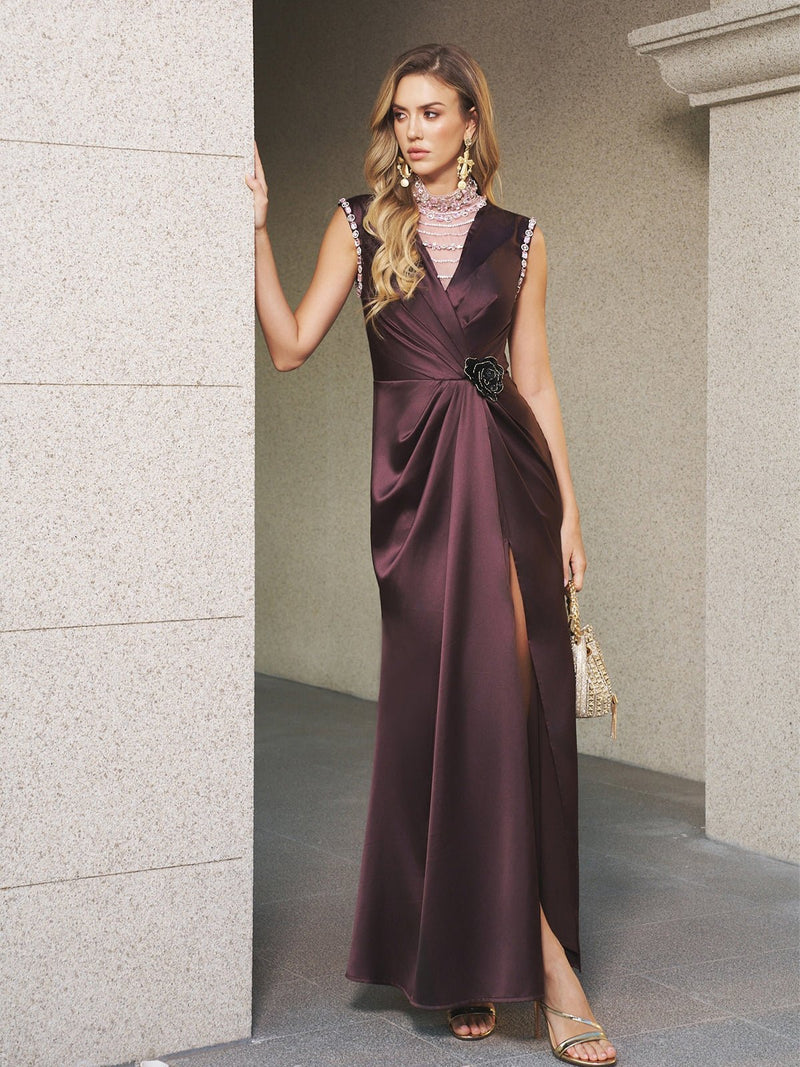 Woman wearing the Social Queen Beaded Satin Floral Appliqué Dress, a sleeveless gown in dark purple with a high slit and fishtail silhouette, standing by a stone wall and holding a small handbag.