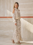 A woman in the Social Queen Beaded Sequin Cold Shoulder Top and high-heeled sandals stands in a modern, beige-toned interior.