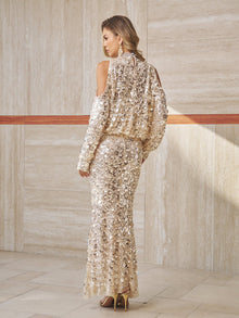 A woman stands indoors wearing the Social Queen Beaded Sequin Cold Shoulder Top, styled with high heels and facing away from the camera against a beige tiled wall.