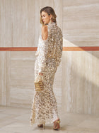 A woman in the Social Queen Beaded Sequin Mermaid Elastic Skirt with cold shoulder sleeves stands indoors, holding a matching gold clutch and wearing gold heels.