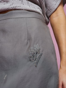 Close-up of a person wearing the Social Queen Beaded Silk Organza Midi Skirt, featuring beaded floral embroidery near the hip and a midi silhouette, paired with a textured gray top; light pink background.