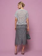 A woman faces away in a sheer light gray crocheted top with beaded embroidery, the Social Queen Beaded Silk Organza Midi Skirt featuring a back slit, heeled sandals, and a textured handbag against a pink background.