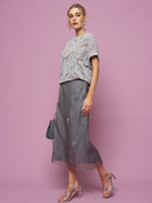 Woman stands against a pink background wearing a light gray knit top, Social Queen's Beaded Silk Organza Midi Skirt, silver heeled sandals, and holding a small gray handbag.