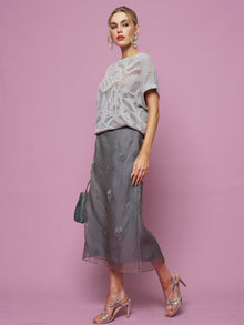 Woman stands against a pink background wearing a light gray knit top, Social Queen's Beaded Silk Organza Midi Skirt, silver heeled sandals, and holding a small gray handbag.