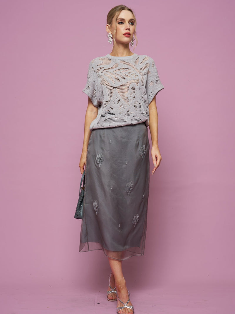 A woman stands against a pink background wearing a light gray crocheted top, silver sandals, dangling earrings, and holding a small handbag. She showcases the Social Queen Beaded Silk Organza Midi Skirt.