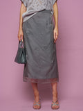 A person in Social Queen’s Beaded Silk Organza Midi Skirt, a grey knit top, and silver sandals stands against a pink background, holding a grey beaded handbag.