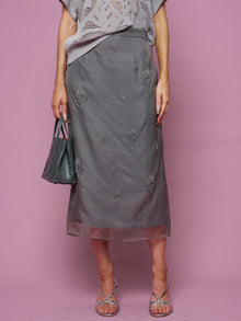 A person in Social Queen’s Beaded Silk Organza Midi Skirt, a grey knit top, and silver sandals stands against a pink background, holding a grey beaded handbag.