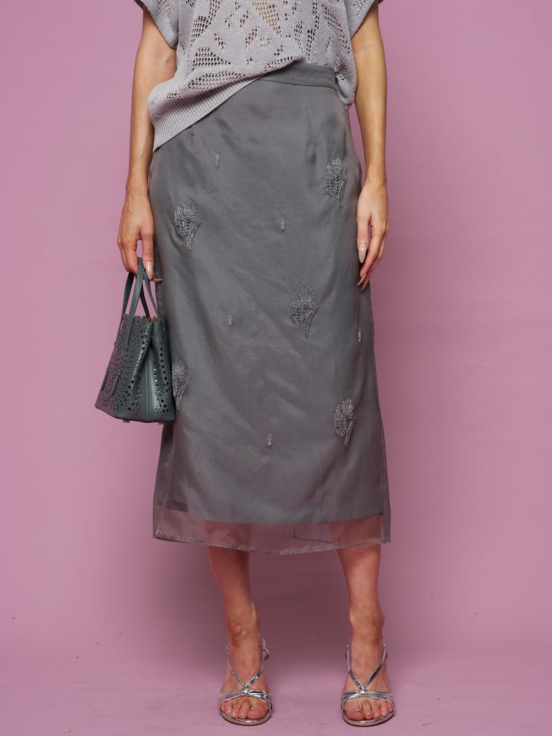 A person in Social Queen’s Beaded Silk Organza Midi Skirt, a grey knit top, and silver sandals stands against a pink background, holding a grey beaded handbag.
