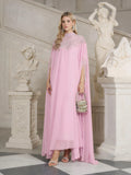A woman in the Social Queen Beaded Tulle Caftan Cape Dress stands indoors by white classical sculptures, holding a small embellished handbag and wearing heeled sandals.