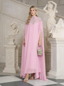 A woman in the Social Queen Beaded Tulle Caftan Cape Dress stands indoors by white classical sculptures, holding a small embellished handbag and wearing heeled sandals.