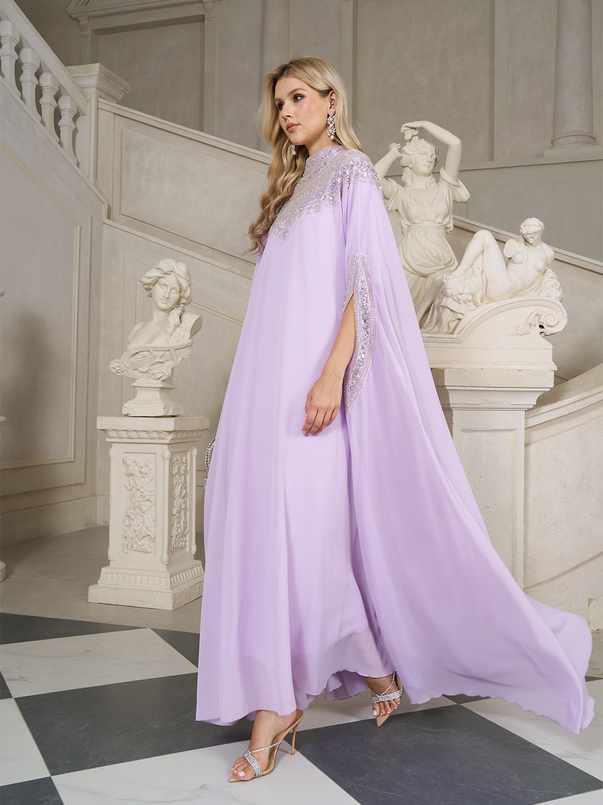 A woman in a Beaded Tulle Caftan Cape Dress by Social Queen and heels walks indoors past classical sculptures and marble stairs.
