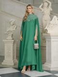 A woman in a Social Queen Beaded Tulle Caftan Cape Dress holds a rhinestone handbag, surrounded by white classical statues and ornate architecture.