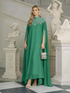 A woman in a Social Queen Beaded Tulle Caftan Cape Dress holds a rhinestone handbag, surrounded by white classical statues and ornate architecture.