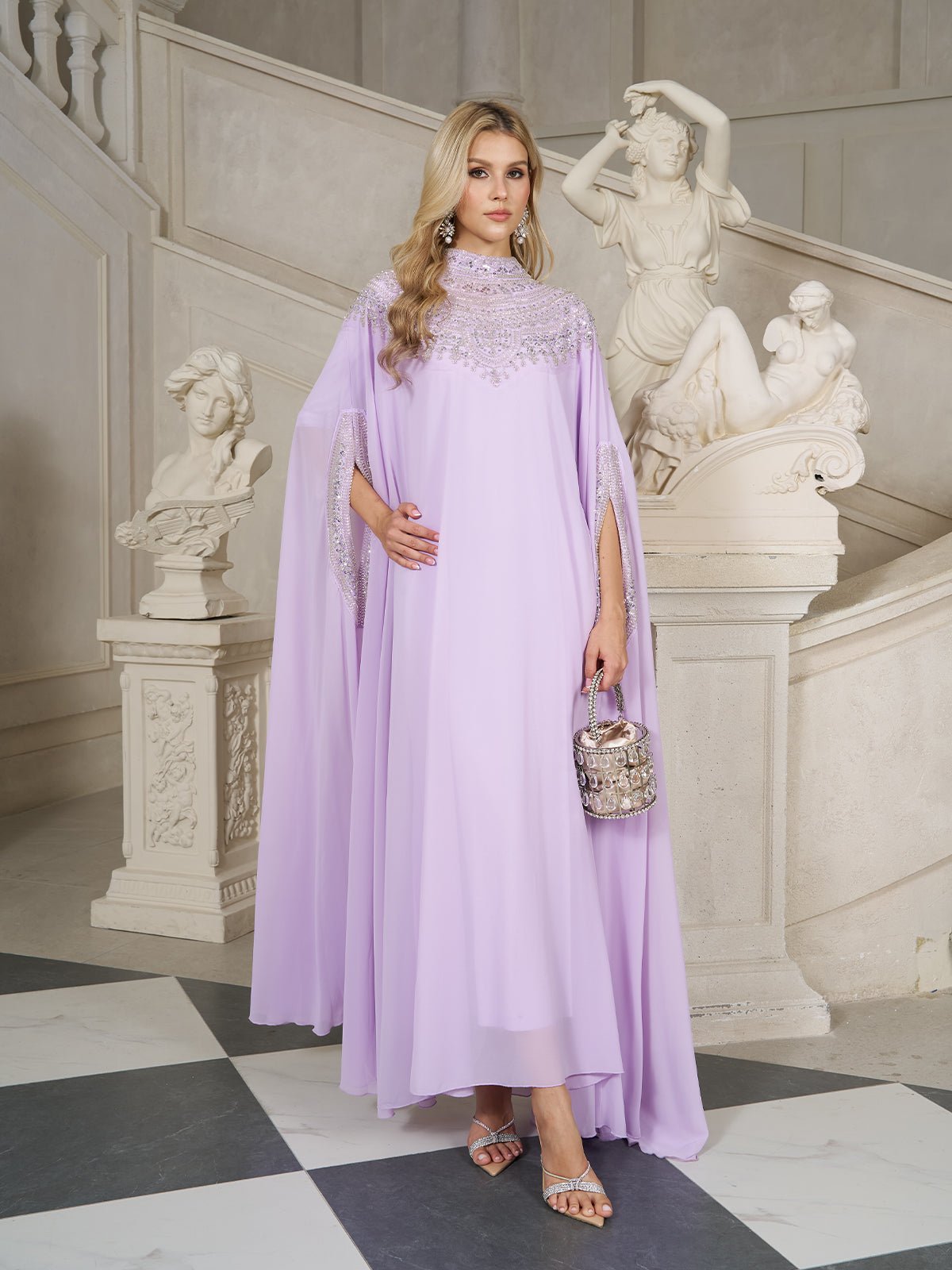 A woman in the Social Queen Beaded Tulle Caftan Cape Dress stands indoors near ornate statues and a white staircase, holding a small embellished purse.