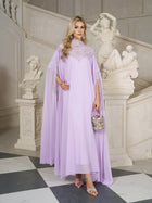 A woman in the Social Queen Beaded Tulle Caftan Cape Dress stands indoors near ornate statues and a white staircase, holding a small embellished purse.