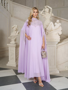 A woman in the Social Queen Beaded Tulle Caftan Cape Dress stands indoors near ornate statues and a white staircase, holding a small embellished purse.
