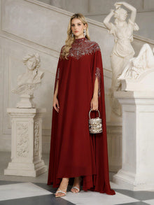A woman in a Social Queen Beaded Tulle Caftan Cape Dress stands indoors by classical statues, holding a small metallic handbag and wearing statement earrings.