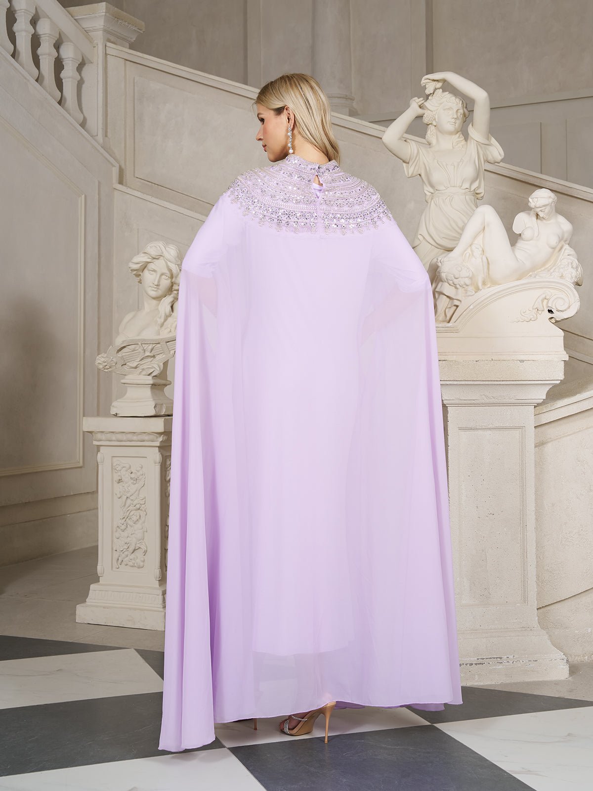 A blonde woman stands in an elegant room near white classical statues and a staircase, wearing the Social Queen Beaded Tulle Caftan Cape Dress, facing away from the camera.