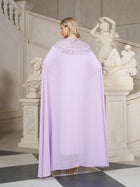 A blonde woman stands in an elegant room near white classical statues and a staircase, wearing the Social Queen Beaded Tulle Caftan Cape Dress, facing away from the camera.