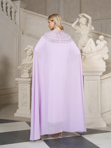 A blonde woman stands in an elegant room near white classical statues and a staircase, wearing the Social Queen Beaded Tulle Caftan Cape Dress, facing away from the camera.