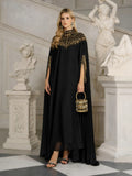 A woman stands in an ornate room wearing the Social Queen Beaded Tulle Caftan Cape Dress, holding a small decorated handbag and surrounded by classical statues.